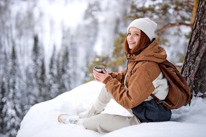 Smiling redhead woman sits on snow on top of mountains relaxing, drinking hot tea from thermos.