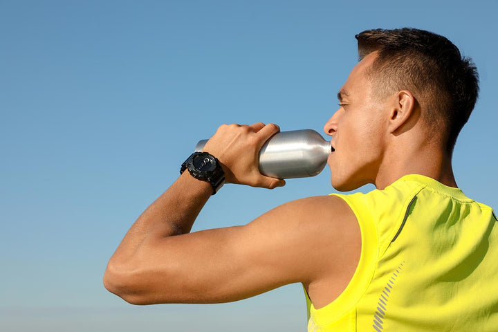 Young athlete drinks from water bottle against blue sky on sunny day.